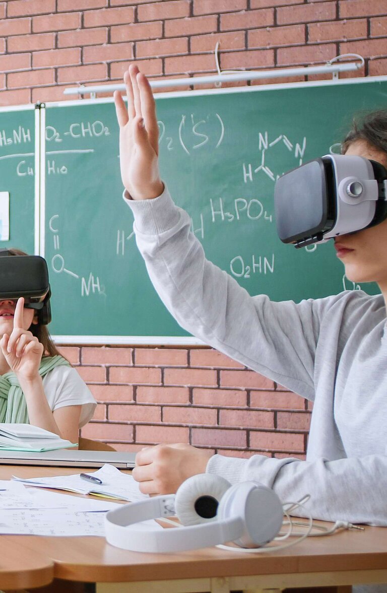 Sechs Schüler:innen sitzen mit VR-Brillen an einem Tisch in einem Klassenzimmer mit grünem Chemie-Tafelbild. Eine Person hebt die Hand, während eine Lehrkraft im Hintergrund mit einem Tablet interagiert. Auf dem Tisch liegen Notizbücher und Stifte.