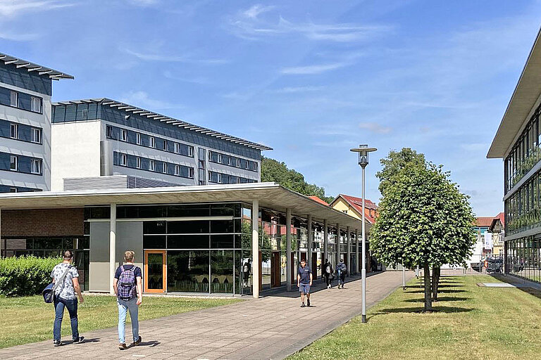 Campusgelände mit mehreren modernen Gebäuden, darunter die Hochschule Schmalkalden im Hintergrund. Menschen gehen auf einem gepflasterten Weg zwischen gepflegten Grünflächen und Bäumen entlang.