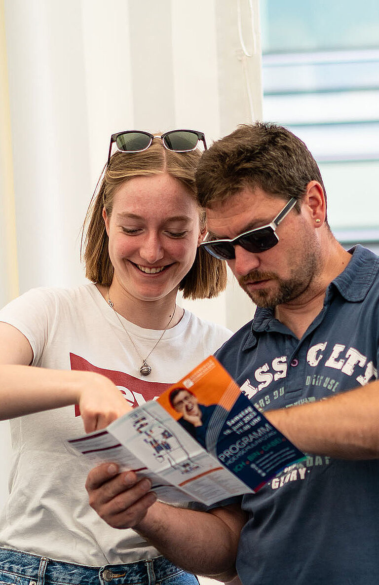 Zwei Personen stehen nebeneinander und betrachten gemeinsam ein Faltblatt. Die linke Person trägt ein weißes T-Shirt und zeigt lächelnd auf das Papier, die rechte Person trägt ein blaues Shirt und Sonnenbrille. Im Hintergrund moderne Architektur mit Glasfassade.