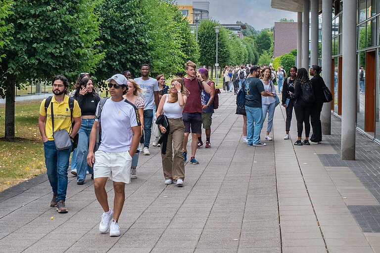 Studierende gehen auf einem belebten Campusweg entlang, flankiert von modernen Gebäuden mit großen Fenstern und grünen Bäumen. Einige tragen Rucksäcke, andere unterhalten sich – typische Szene eines aktiven Hochschultages.