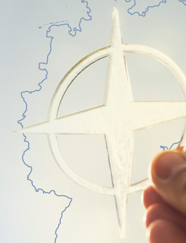 Hand hält ein transparentes NATO-Emblem aus milchigem Material vor einer dünn gezeichneten Umrisskarte von Deutschland; im Hintergrund verläuft der Himmel in einem sanften Blau-Hell-Verlauf.