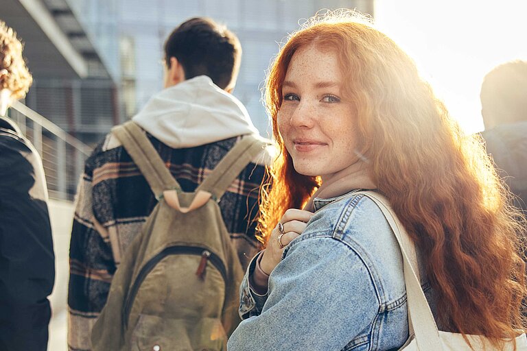 Fünf junge Menschen gehen gemeinsam durch ein urbanes Hochschulumfeld. Eine Person mit roten Haaren und Jeansjacke blickt lächelnd zurück zur Kamera, während die anderen mit Rucksäcken vorausgehen. Im Hintergrund moderne Gebäude und Sonnenlicht.
