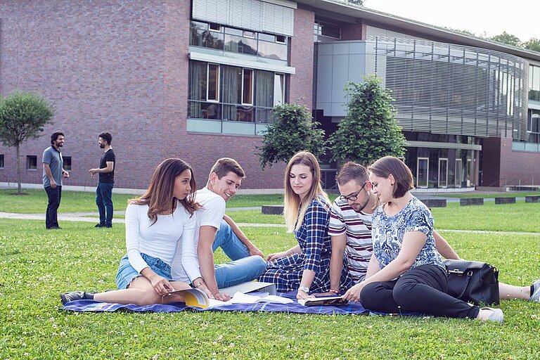„Sechs junge Erwachsene sitzen auf einer Decke auf einer Wiese vor einem modernen Backsteingebäude mit großen Fenstern. Sie sind in eine Lerngruppe vertieft, mit Notizbüchern und Unterlagen vor sich. Im Hintergrund gehen zwei weitere Personen am Gebäude entlang.