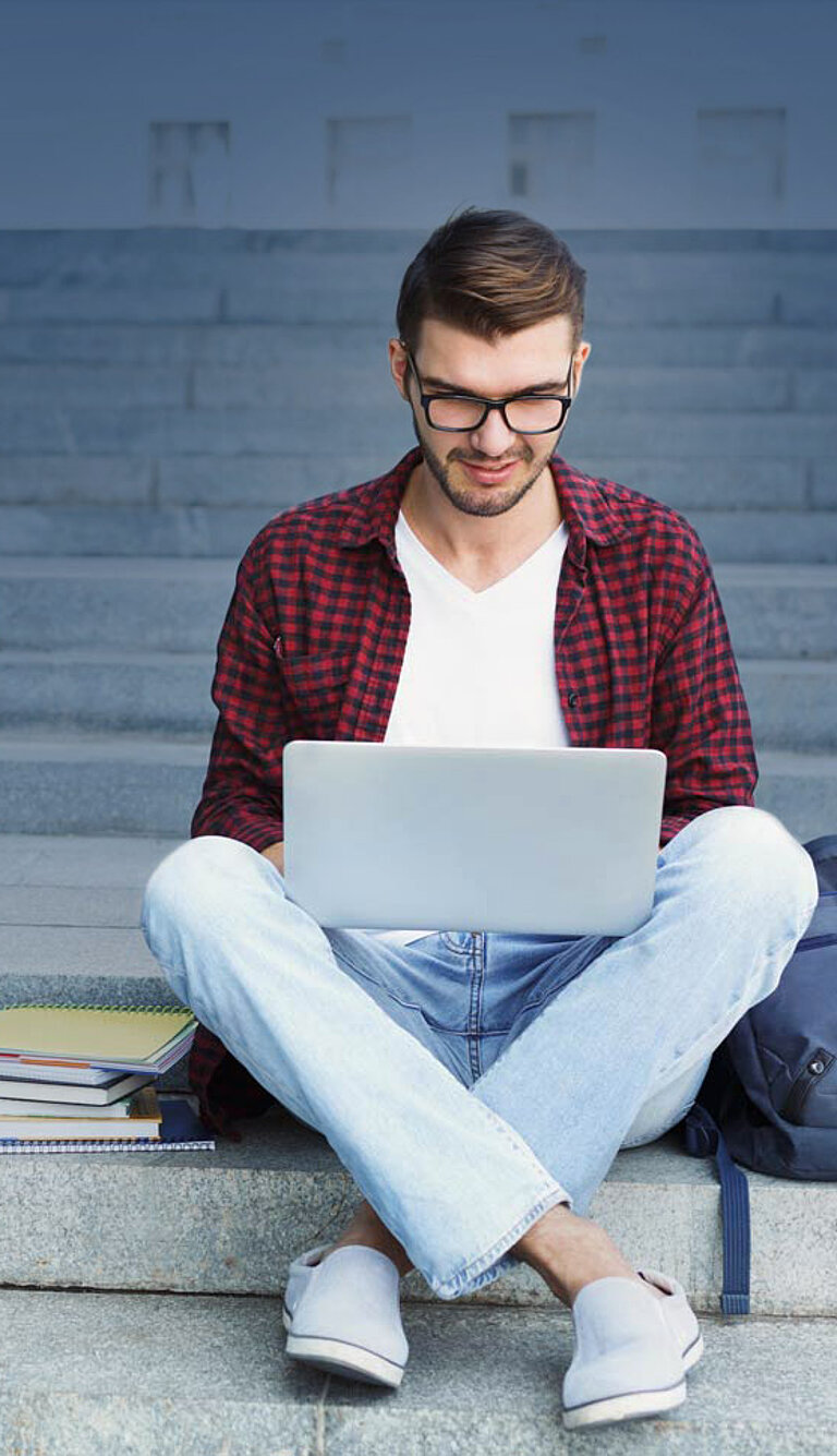 Person mit Brille und kariertem Hemd sitzt auf Außentreppen vor einem Hochschulgebäude, arbeitet am Laptop. Daneben liegen ein blauer Rucksack und Bücher – Szene aus dem studentischen Alltag.