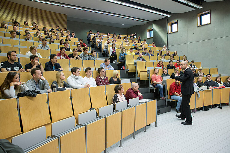 Studierende sitzen in einem modernen Hörsaal mit gestuften Holzstühlen und Betonwänden. Ein Dozent im Anzug steht vorne und spricht, während die Studierenden aufmerksam zuhören und mitschreiben.